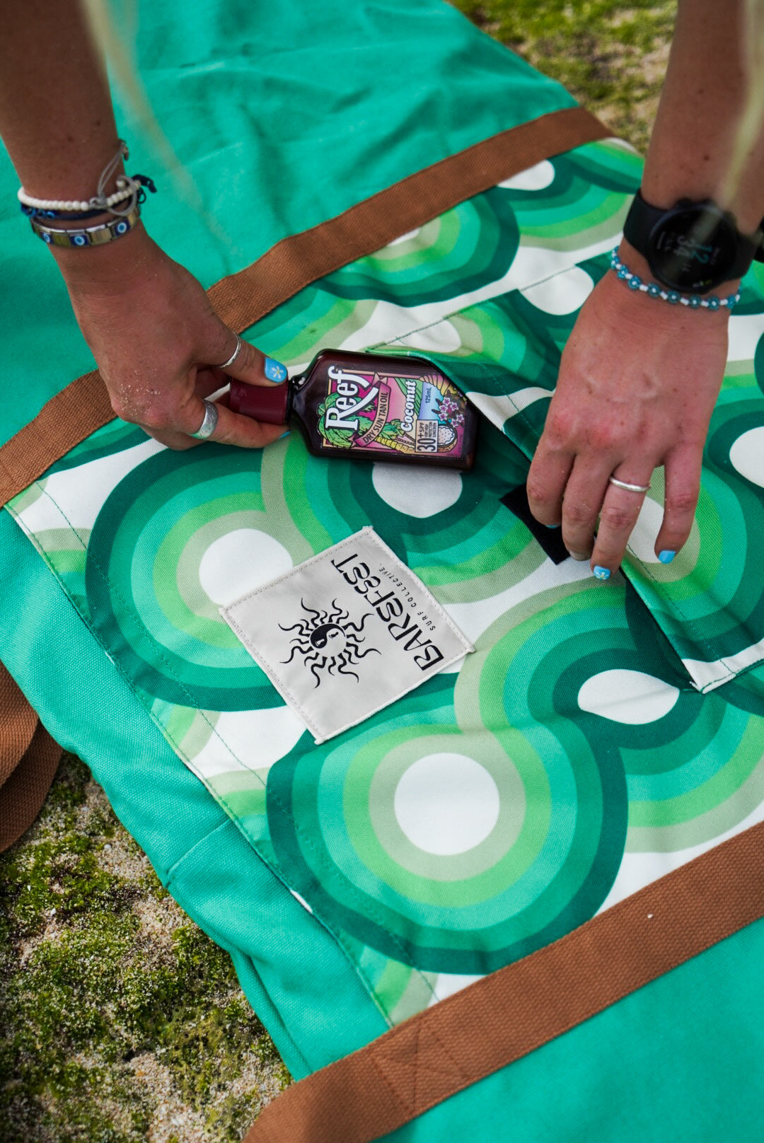 This image features a person standing on a sandy beach with a surfboard cover designed to resemble a lush, green aquatic scene, capturing the essence of the coastline. The surfboard cover, in shades of green, is patterned to mimic the natural hues found along the South Australian coast.