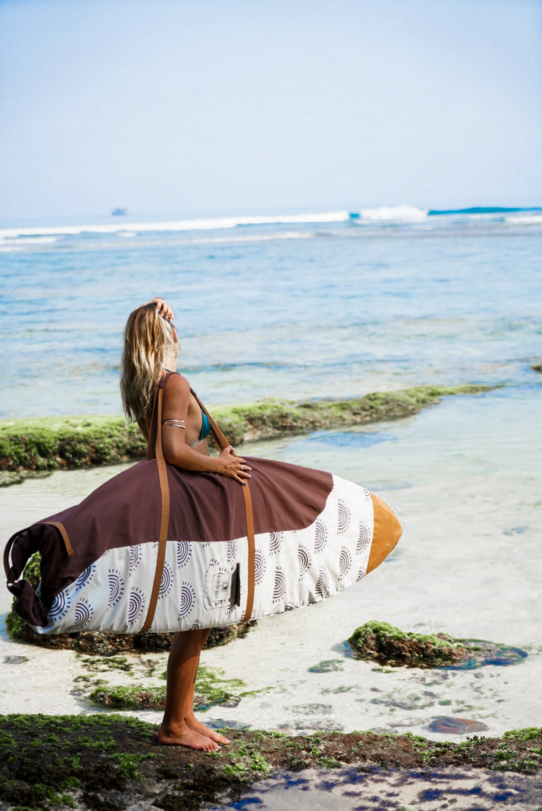 This image features a person carrying a surfboard, which is covered with a mid-length canvas surfboard cover. The cover displays a unique sandy brown tone, reminiscent of the South Australian coastlines.