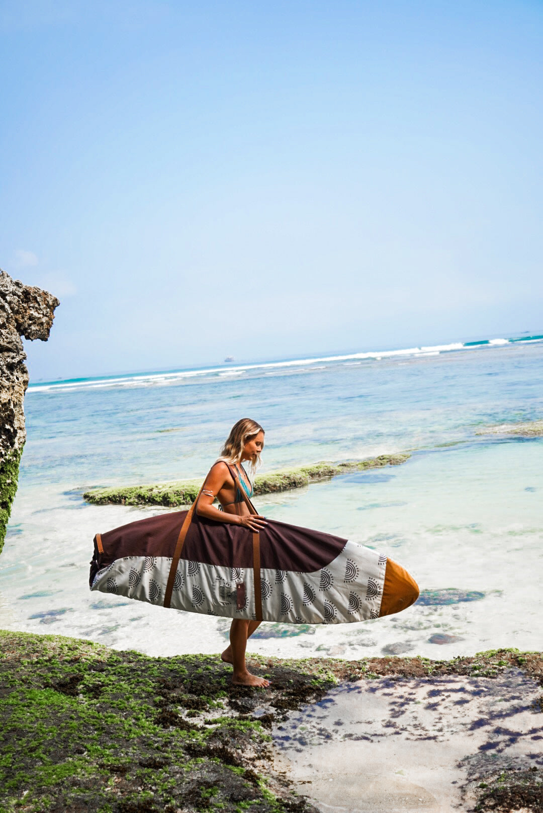 This image features a person carrying a surfboard, which is covered with a mid-length canvas surfboard cover. The cover displays a unique sandy brown tone, reminiscent of the South Australian coastlines.