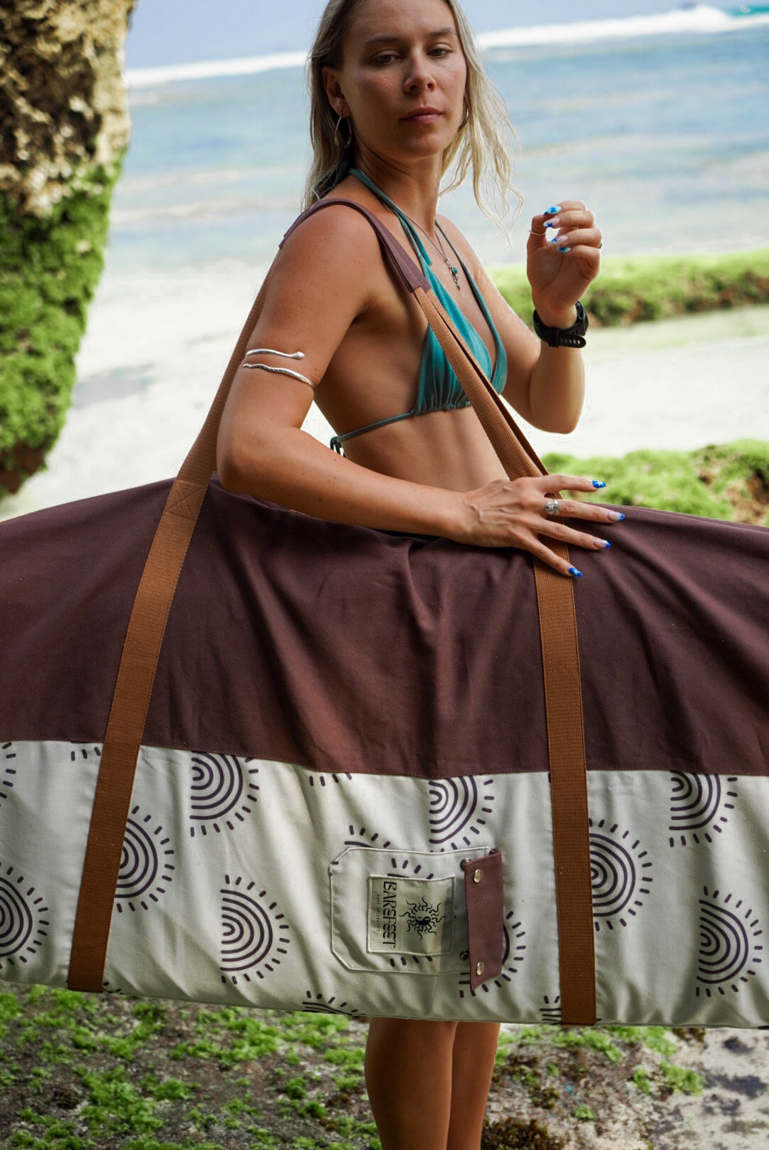 This image features a person carrying a surfboard, which is covered with a mid-length canvas surfboard cover. The cover displays a unique sandy brown tone, reminiscent of the South Australian coastlines.