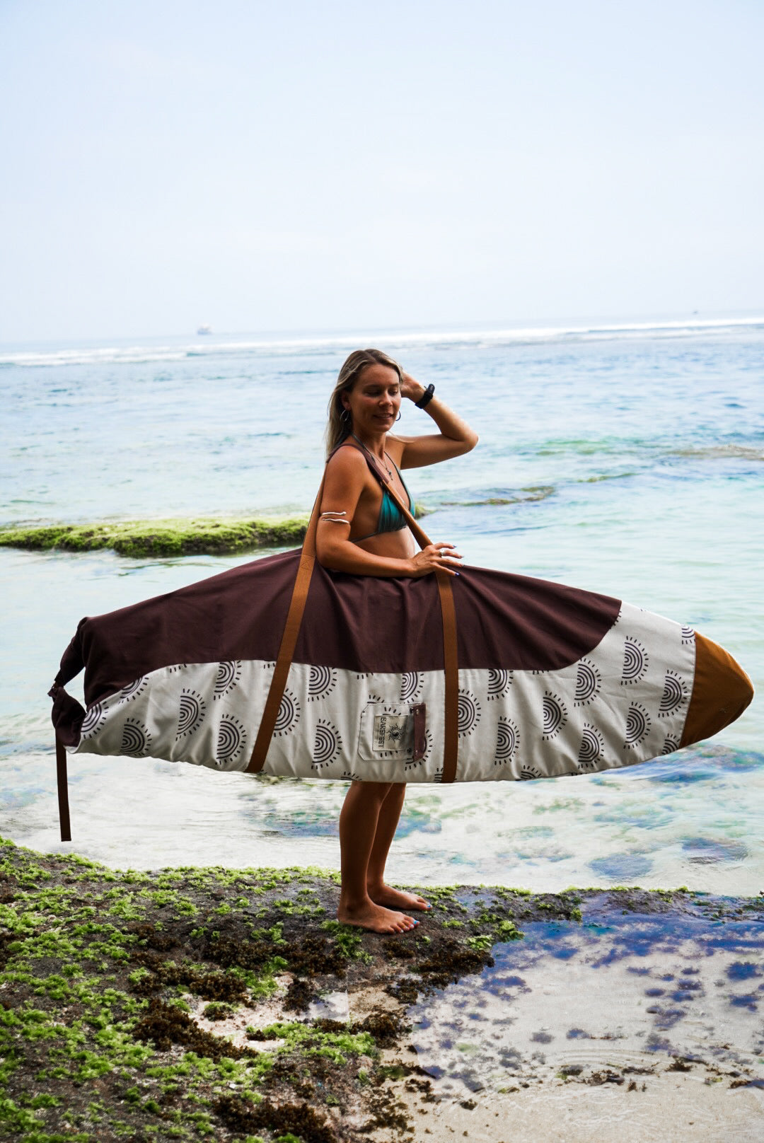 This image features a person carrying a surfboard, which is covered with a mid-length canvas surfboard cover. The cover displays a unique sandy brown tone, reminiscent of the South Australian coastlines.