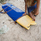 This image features a person standing on a sandy beach with the ocean in the background, holding a mid-length canvas surfboard cover. The cover displays a striking ocean blue color with a white geometric pattern near the bottom.