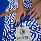 This image features a person standing on a sandy beach with the ocean in the background, holding a mid-length canvas surfboard cover. The cover displays a striking ocean blue color with a white geometric pattern near the bottom.