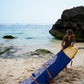 This image features a person standing on a sandy beach with the ocean in the background, holding a mid-length canvas surfboard cover. The cover displays a striking ocean blue color with a white geometric pattern near the bottom.