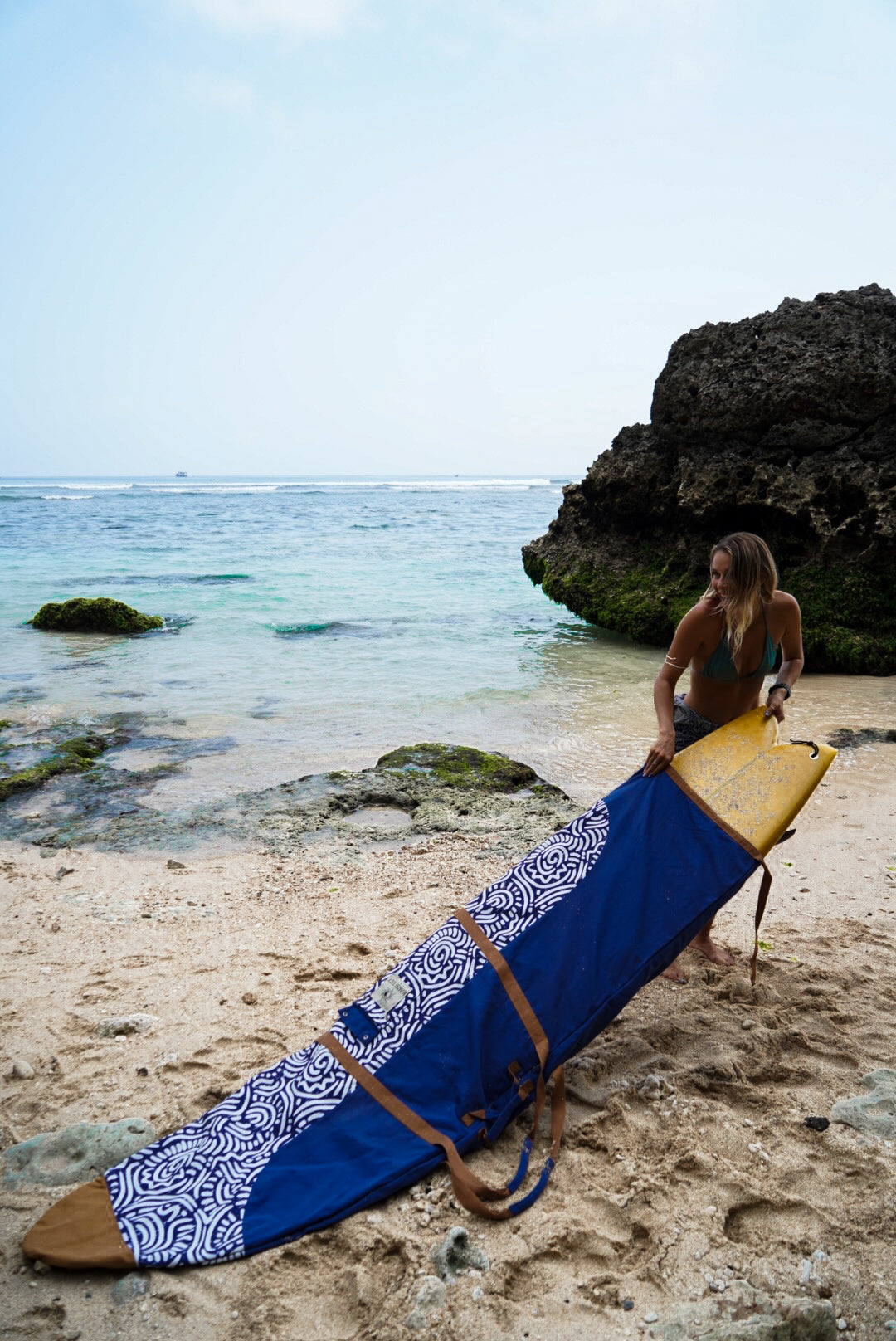 This image features a person standing on a sandy beach with the ocean in the background, holding a mid-length canvas surfboard cover. The cover displays a striking ocean blue color with a white geometric pattern near the bottom.