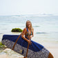 This image features a person standing on a sandy beach with the ocean in the background, holding a mid-length canvas surfboard cover. The cover displays a striking ocean blue color with a white geometric pattern near the bottom.