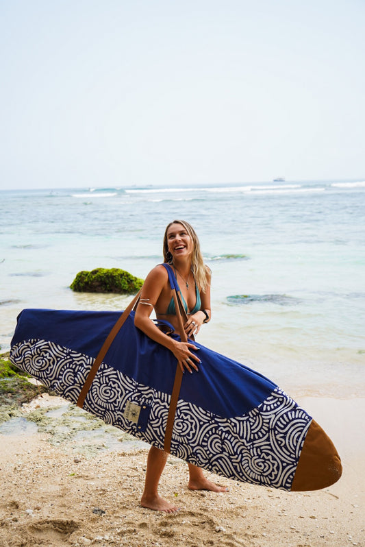 This image features a person standing on a sandy beach with the ocean in the background, holding a mid-length canvas surfboard cover. The cover displays a striking ocean blue color with a white geometric pattern near the bottom.