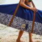 This image features a person standing on a sandy beach with the ocean in the background, holding a mid-length canvas surfboard cover. The cover displays a striking ocean blue color with a white geometric pattern near the bottom.
