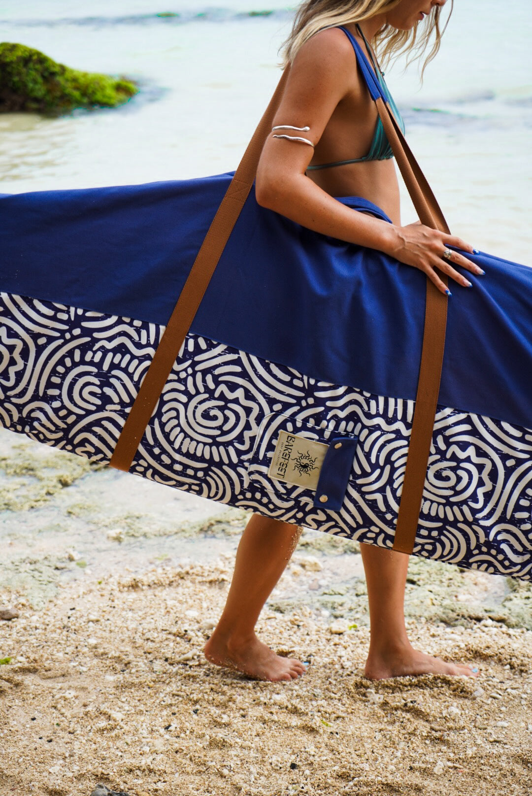 This image features a person standing on a sandy beach with the ocean in the background, holding a mid-length canvas surfboard cover. The cover displays a striking ocean blue color with a white geometric pattern near the bottom.
