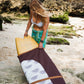 This image features a person carrying a surfboard, which is covered with a mid-length canvas surfboard cover. The cover displays a unique sandy brown tone, reminiscent of the South Australian coastlines.
