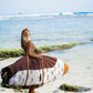 This image features a person carrying a surfboard, which is covered with a mid-length canvas surfboard cover. The cover displays a unique sandy brown tone, reminiscent of the South Australian coastlines.