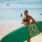 This image features a person standing on a sandy beach with a surfboard cover designed to resemble a lush, green aquatic scene, capturing the essence of the coastline. The surfboard cover, in shades of green, is patterned to mimic the natural hues found along the South Australian coast.