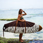 This image features a person carrying a surfboard, which is covered with a mid-length canvas surfboard cover. The cover displays a unique sandy brown tone, reminiscent of the South Australian coastlines.