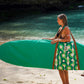 This image features a person standing on a sandy beach with a surfboard cover designed to resemble a lush, green aquatic scene, capturing the essence of the coastline. The surfboard cover, in shades of green, is patterned to mimic the natural hues found along the South Australian coast.