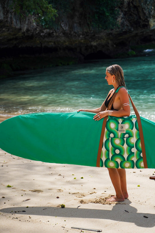 This image features a person standing on a sandy beach with a surfboard cover designed to resemble a lush, green aquatic scene, capturing the essence of the coastline. The surfboard cover, in shades of green, is patterned to mimic the natural hues found along the South Australian coast.